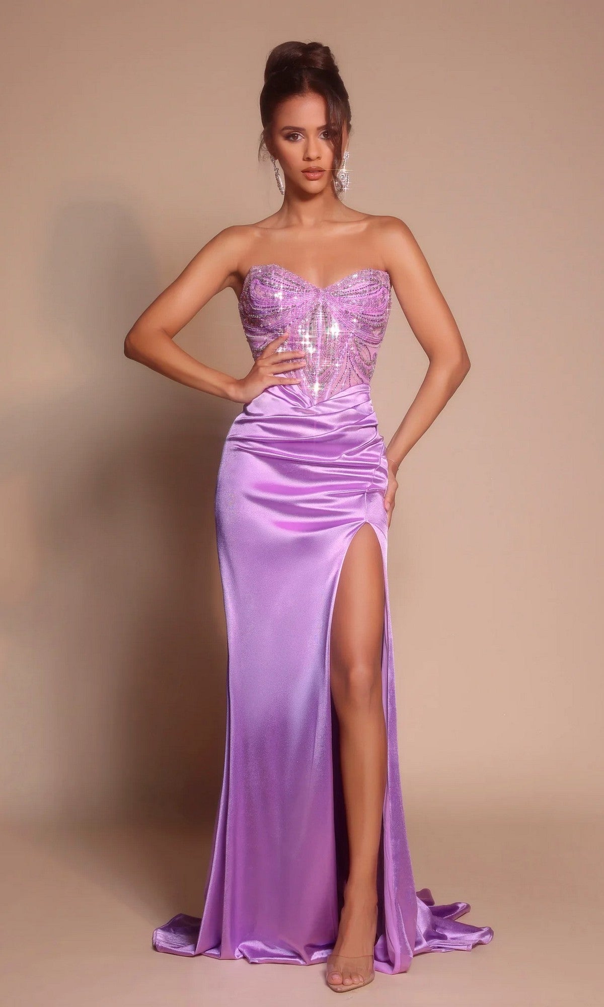 A woman poses in the lilac Portia and Scarlett XO065 long formal dress, featuring a strapless embellished corset bodice and thigh-high slit, against a plain beige background.